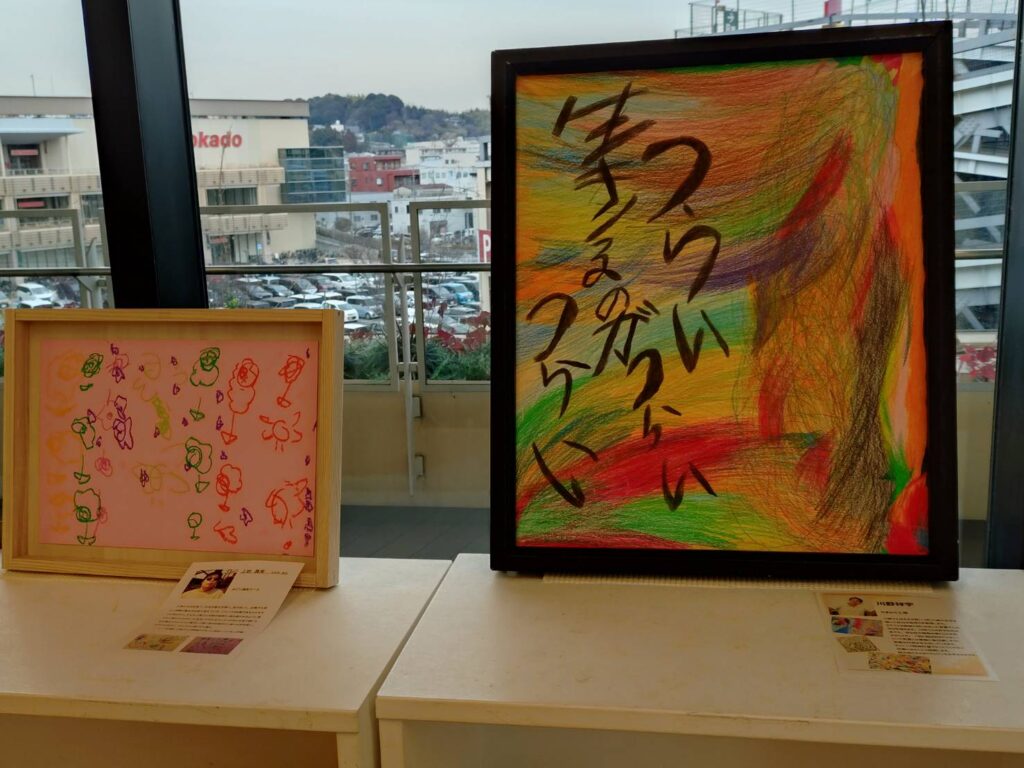 様々な色が渦を巻くように描かれた背景の上に「つらい　生きるのがつらい　つらい」と書かれた絵画作品