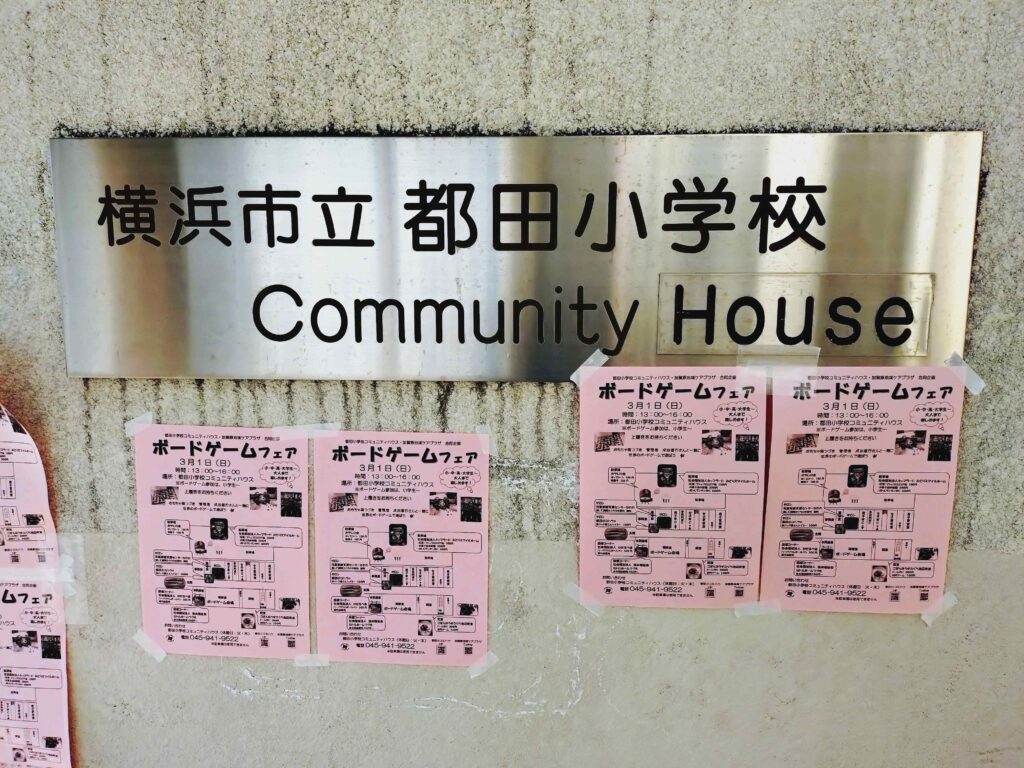都田小学校コミュニティハウス看板の周辺にボードゲームフェアのチラシが複数枚貼られている