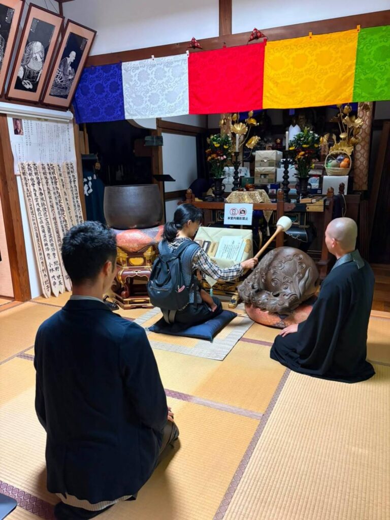 住職の指示を受けて木魚を叩くなつみ編集長と、見守るさかす編集長